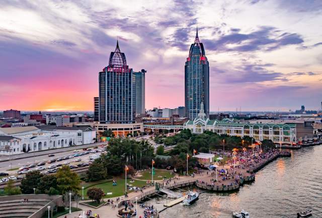 A vibrant sunset over Downtown Mobile skyline, featuring tall buildings, a bustling waterfront, and a lively crowd below.