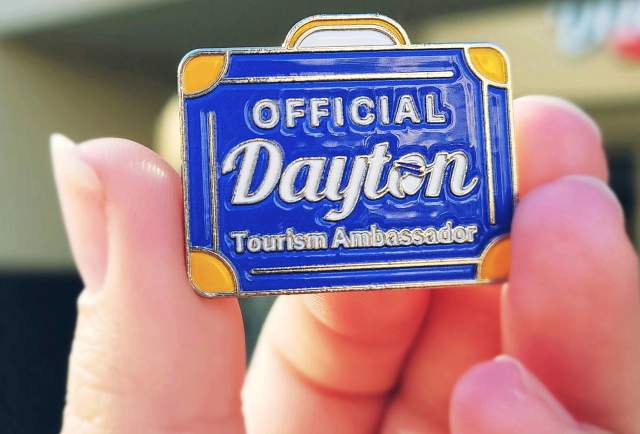 a close-up of a hand holding a blue pin with the text official dayton tourism ambassador on it
