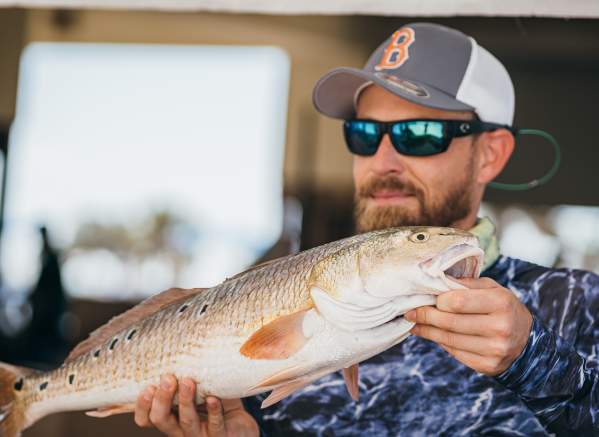 A smiling man in a cap and sunglasses holds up a speckled trout