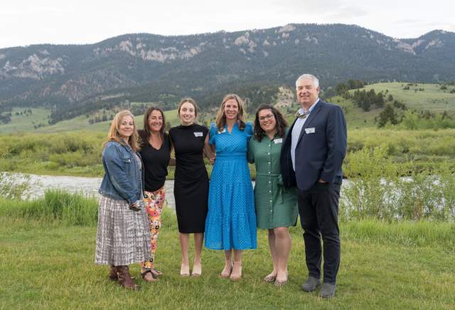 A group of six people stands on a grassy area by a river, with mountains in the background. The scene is set in a natural, outdoor environment.
