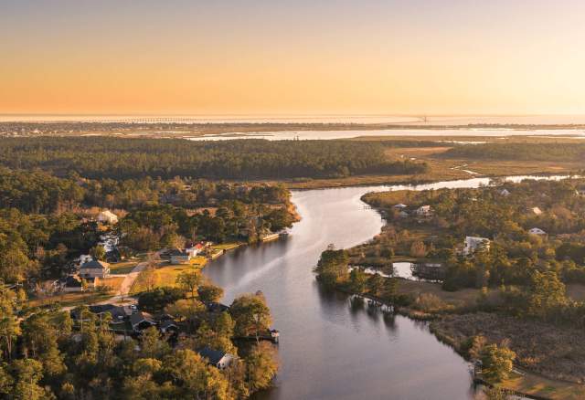 Aerial sunset view of a winding Slidell waterway bordered by homes, marshes and trees, with Lake Pontchartrain stretching across the horizon on the Fall/Winter 2026–27 cover of The Northshore.
