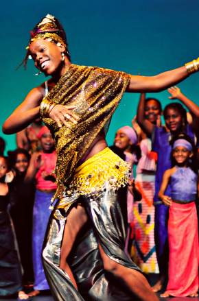 A Kirikou performer at Orlando Fringe festival in vibrant, traditional attire performs energetically on stage, surrounded by a lively group of children and adults dressed in colorful clothing, celebrating in a joyful and cultural setting.