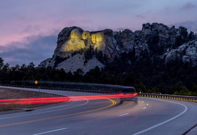 Mount Rushmore, lit up by yellow floodlights at dusk, sits in the background as a vehicle traveling down the highway is trailed by a long line of red taillights, created by a long-exposure photo.