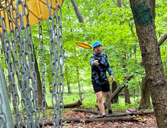Man throws disc golf frisbee towards camea, which sits behind the hole in heavily wooded and shaded area