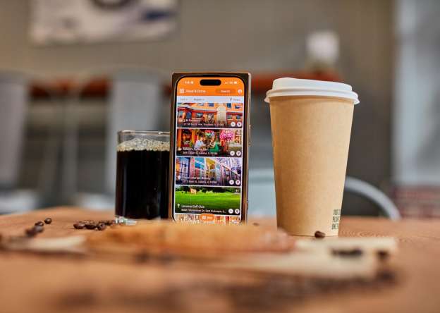 Two cups of coffee, one in a to-go mug, and a cellphone with the Galena Country App pulled up. All set on a table covered in coffee beans and a cookie.