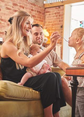 A family sits on a couch at a coffee shop while a mom feeds a child a bite of her food.