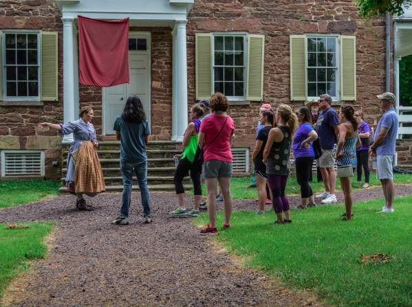 A group of visitors listening to a woman dressed in historic attire outside of a brick building