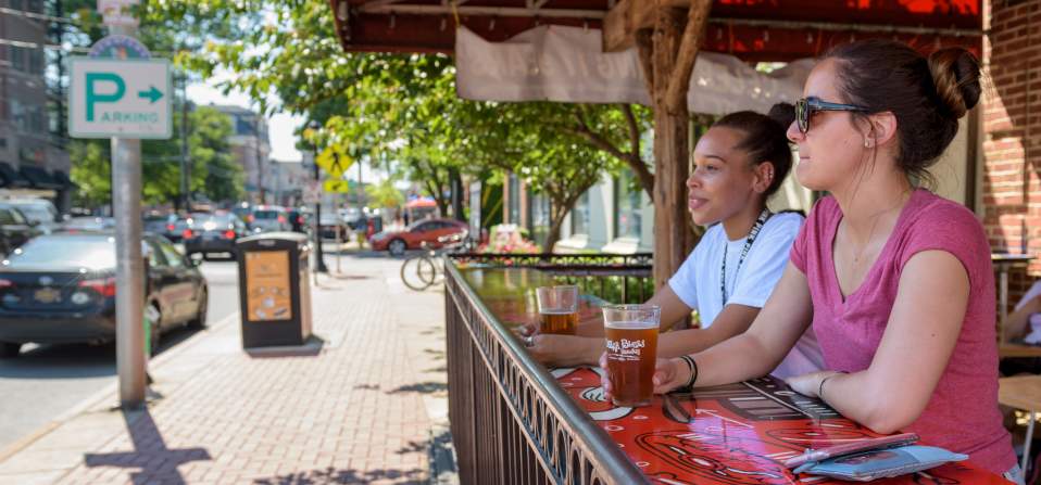 Two people sitting at a colorful outdoor bar table with drinks, beside a sidewalk on Main Street, Newark, Delaware, with cars and parking signs.
