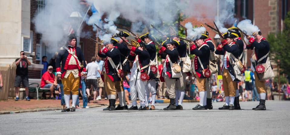 A group of historical reenactors in colonial military uniforms fires muskets, creating clouds of smoke during a lively outdoor event.