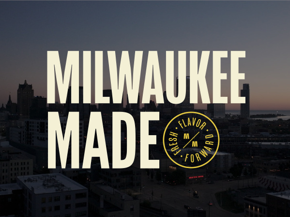 Aerial view of Milwaukee's downtown skyline at dusk with tall buildings silhouetted against a gradient sky. Large cream-colored text reads 'MILWAUKEE MADE' overlaid on the cityscape, with a circular yellow badge logo containing 'FRESH FLAVOR FORWARD' and stylized 'MM' letters positioned to the right of 'MADE'.