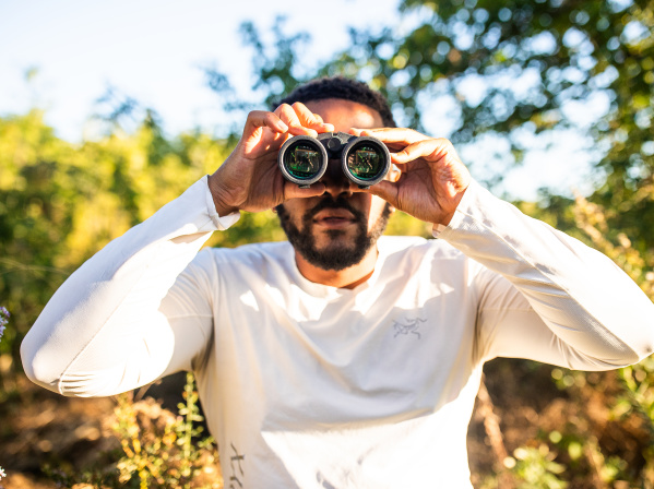 A man in a white long-sleeved shirt holds binoculars up to his eyes while standing outdoors in a sunny, tree-filled setting with green foliage and dappled sunlight in the background.