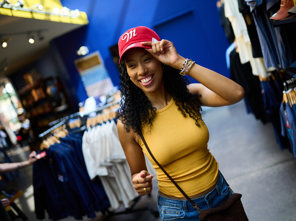 Smiling shopper trying on a red Miller-branded baseball cap inside the Miller Brewery gift shop, surrounded by racks of apparel and branded merchandise.
