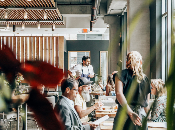 Diners enjoy a meal inside a bright, modern Milwaukee restaurant with large windows, warm lighting, and a relaxed, welcoming atmosphere as a chef engages with guests.