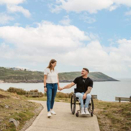 A man in a wheelchair holding hands with a standing woman on a section of the coast path with the sea in the background.