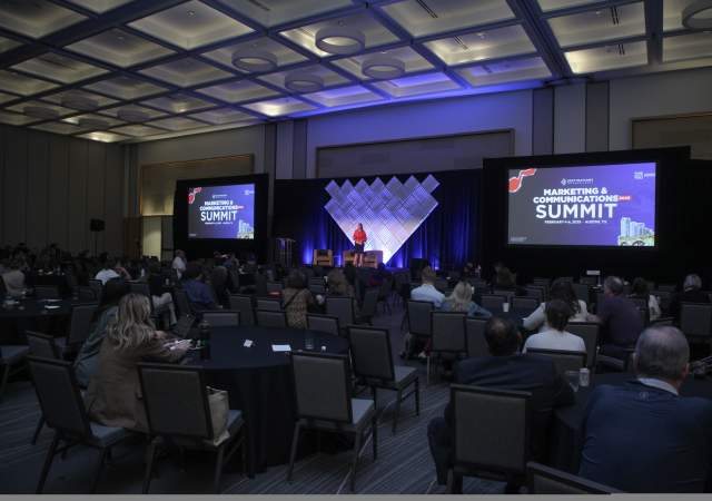 Woman standing on stage at DI Marketing & Communications Summit 2025, giving a welcome message to attendees in a ballroom.