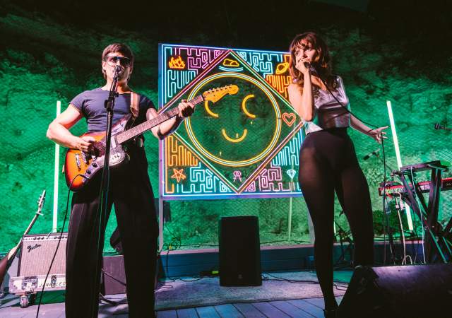 Man playing guitar and woman singing into a microphone on a stage backlit by a neon smiley face.