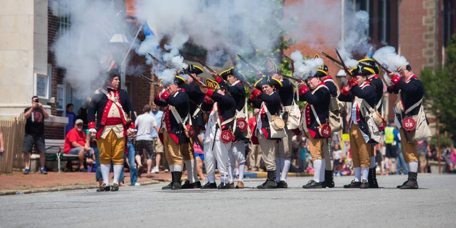 A group of historical reenactors in colonial military uniforms fires muskets, creating clouds of smoke during a lively outdoor event.
