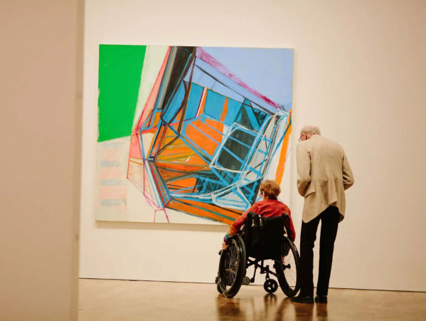 Two elderly people, one in a wheelchair, admire a large, colorful abstract painting in a bright art gallery.