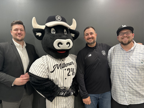 Three men pose for a photo with the Milwaukee Milkmen mascot, a smiling black and white cow wearing a pinstriped baseball uniform and cap, standing in front of a plain dark gray wall.