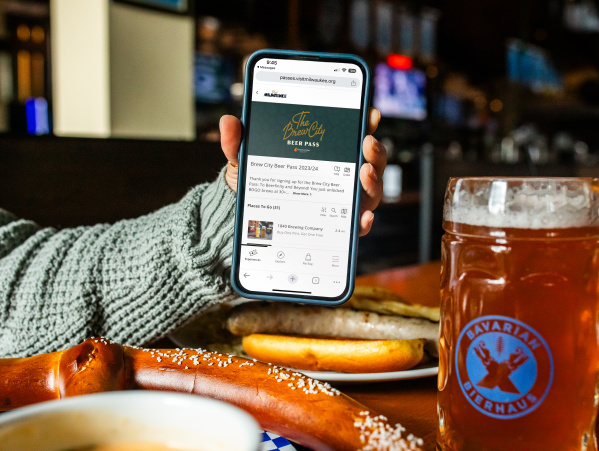 a hand holding up a phone displaying the Brew City Beer Pass over a table with a Bavarian Pretzel and a large pint of beer