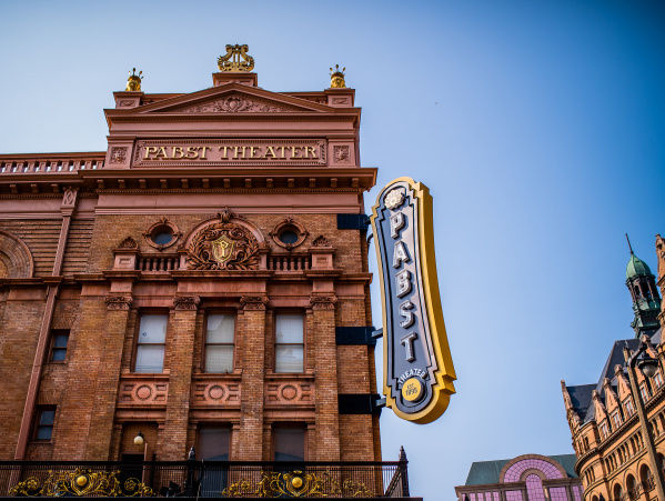 The exterior of the historic Pabst Theater in Milwaukee, featuring its ornate brick façade, gold architectural details, and large vertical “Pabst Theater” marquee sign against a clear blue sky.