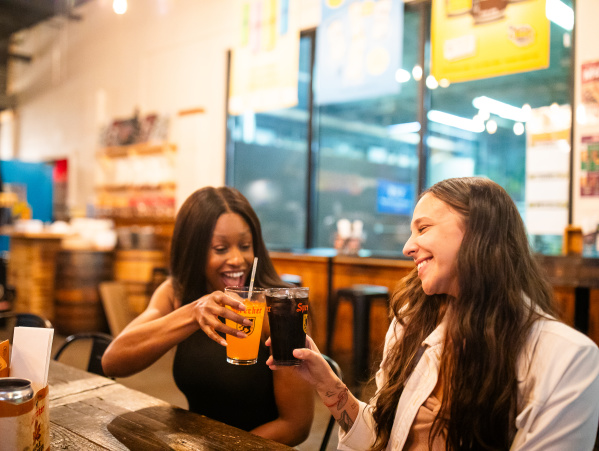 Two women sitting at a wooden table in Sprecher Brewery, smiling and toasting with glasses of beer - one holding a light-colored beer and the other a dark beer. The setting appears warm and friendly with restaurant lighting and blurred background showing other seating areas and windows.