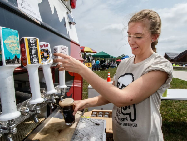 A woman pours a dark beer from a tap at an outdoor beer festival, smiling as she works at a mobile beer truck with colorful tap handles and a crowd visible in the background.