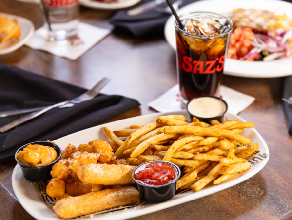 A close-up of a restaurant table featuring a platter of seasoned fries, mozzarella sticks, and dipping sauces. Behind it sits a glass of soda with the restaurant’s logo, and additional plates of food and black napkins are visible in the background.