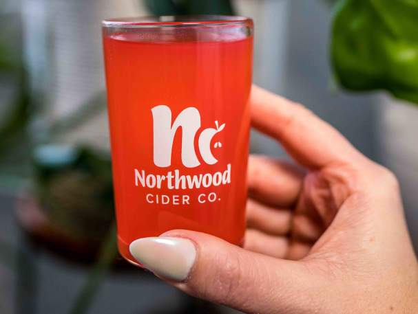 A small glass of a pinky-red liquid is being held up. The glass says Northwood Cider Co.