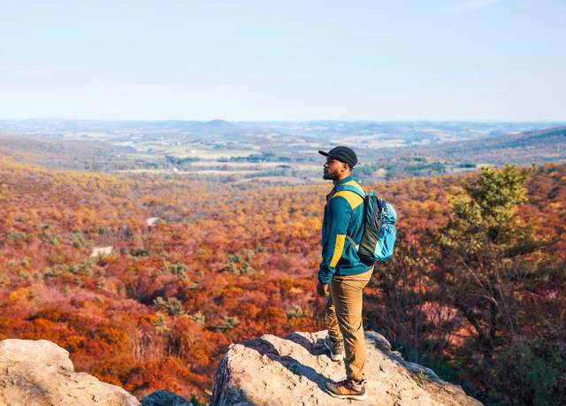 A man with a backpack and hat standing on an overlook looking out at fall foliage
