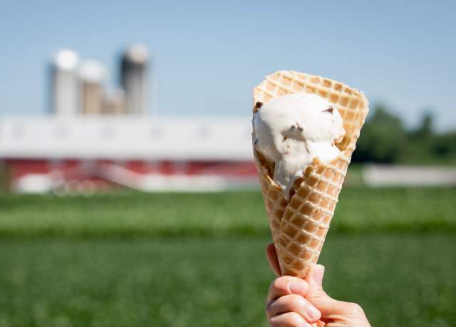 A hand holding up a cone of ice cream in front of a barn