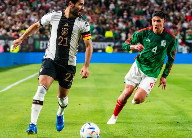 Two men in soccer uniforms kicking a soccer ball on turf at Lincoln Financial Field in Philadelphia during a match with people in the stadium