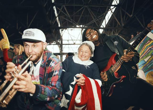 Three people smile and perform music inside a large industrial-style building—one playing trumpet, one playing electric guitar, and a woman in historical dress holding a flag.