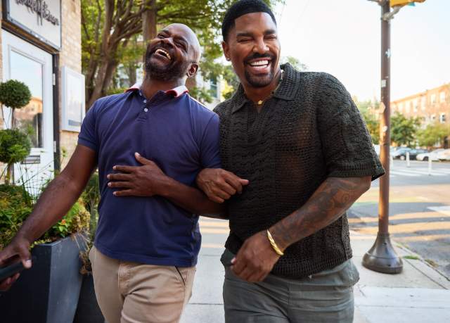 Two African-American men laugh while walking arm in arm down a sidewalk.