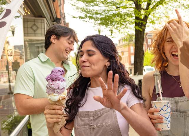 A woman with a large ice cream cone turning away from another woman laughing with a milkshake in her one hand and running her other hand through her hair on a sidewalk on a main street. Two men are giggling and standing around them