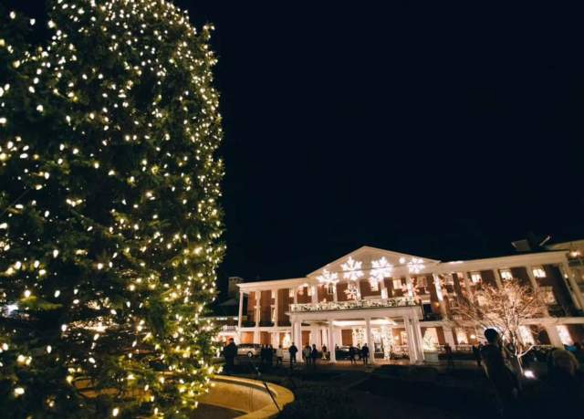 The front entrance of a large mansion, known as Omni Bedford Springs Resort in Bedford, decorated with holiday lights at night with a lit Christmas tree in front.