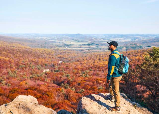 A man with a backpack and hat standing on an overlook looking out at fall foliage