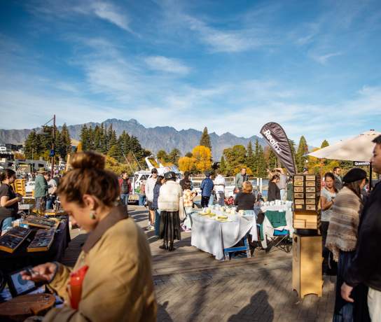 People browsing stalls at Queenstown craft market with autumnal trees and the Remarkables mountain range in the background