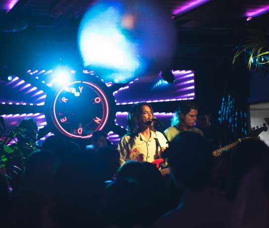 Person singing and playing guitar on a stage with colourful lights and neon sign