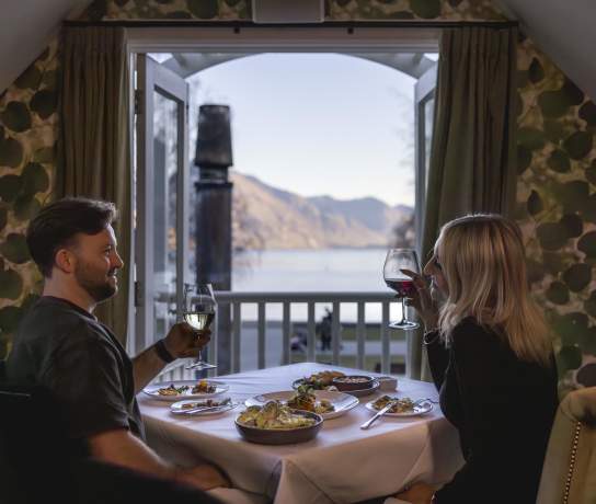 A cosy dinner setting with a couple enjoying wine and a delicious meal, overlooking stunning lake and mountain views through open balcony doors.