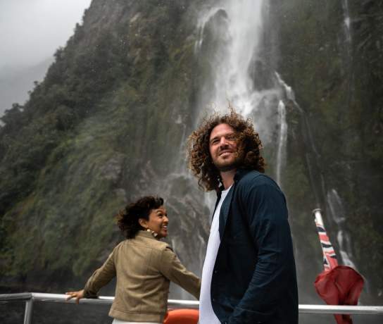 Two people standing on the front of a boat in Milford Sound with a big waterfall in the background
