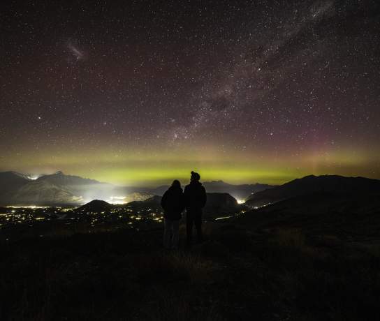People standing on a mountain top with a starry night sky and aroura before them
