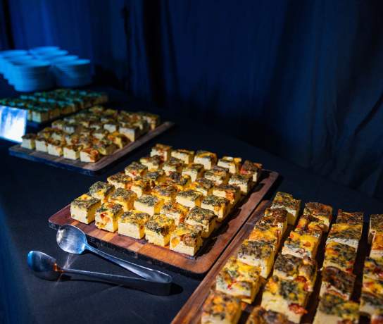 A catering table set up in a dark event room with square quiche's on wooden serving boards