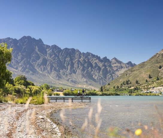 Two people having a rest on a pier looking out over the lake and Remarkables mountains while biking the Frankton Trail