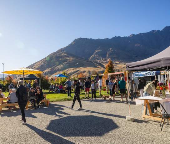 People browsing stalls and relaxing under colourful umbrellas at the Remarkables Market, with mountains in the background on a sunny day.