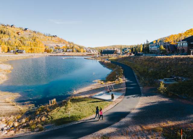 Two people walk on a paved path around a lake surrounded by cabins and autumn trees at Brian Head