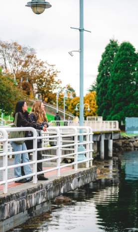 Two people pause along Nanaimo’s waterfront walkway, leaning on a railing and enjoying the view of the calm water and nearby park.