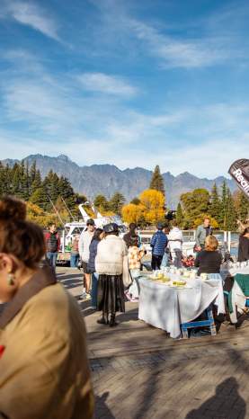 People browsing stalls at Queenstown craft market with autumnal trees and the Remarkables mountain range in the background