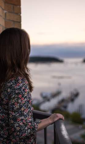 A person stands on a balcony overlooking a harbour with boats and distant islands at sunset.