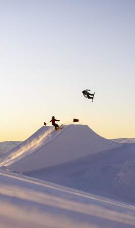 Snowboarders and a skier perform aerial tricks off a jump at Cardrona Alpine Resort at sunrise.